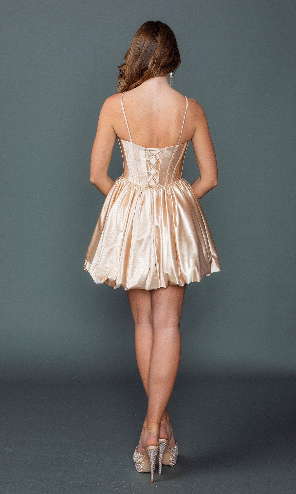 A woman stands with her back to the camera, modeling the Dancing Queen 3435 short homecoming dress—featuring a shiny champagne-colored bubble hem—paired with high-heeled shoes, set against a plain gray background.